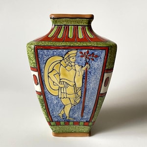 Op de afbeelding: Een keramische vaas met een centraal paneel met een gele figuur die een staf vasthoudt tegen een blauwe gespikkelde achtergrond. De vaas heeft groen getextureerde zijkanten, een rood-groen gestreepte hals en een rood-groen geblokte basis.