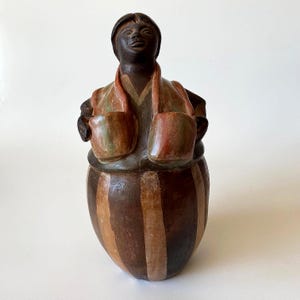 Puede incluir: Figura de cerámica pintada a mano de una persona de tez oscura. La figura lleva un chaleco con dos bolsillos y una falda a rayas. La paleta de colores incluye marrón, rojo y verde.