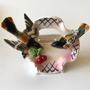 Puede incluir: Una maceta de pared de cerámica con dos pájaros de plumaje negro, blanco y naranja posados en el borde. La maceta es de color crema con un tono rosado, un patrón de rejilla verde oscuro y una hoja verde esculpida con dos cerezas rojas.
