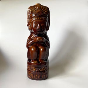 May include: A brown ceramic figurine of a seated figure with a headdress. The figurine has a glossy finish and is inscribed with the word "Cancun" at the base. The figure is holding its hands together in front of its chest.