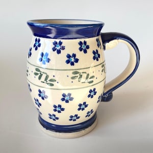 Op de afbeelding: Een keramische mok met een crèmekleurige basis voorzien van een herhalend patroon van kleine blauwe bloemen. Een horizontale band met groene bladmotieven loopt rond het midden. De rand, de voet en het handvat zijn afgewerkt met een donkerblauw glazuur.