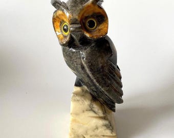 May include: A carved stone owl figurine featuring a dark grey and brown body with prominent yellow eyes. The owl is perched on a light cream-colored stone base with grey veining. The sculpture displays a smooth, polished texture throughout.