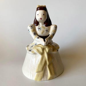 Op de afbeelding: Een keramische bel in de vorm van een figuur met bruin haar, gekleed in een crèmekleurige jurk met kleine blauwe en gele bloemmotieven. De basis van de bel heeft een opvallende gele strik en een zichtbare craquelé glazuurafwerking.