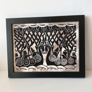 May include: A framed black and white papercut artwork featuring two stylized Viking ships facing each other. The design includes intricate knotwork patterns above the ships, figures with pointed hats, and swirling wave motifs at the bottom edge.