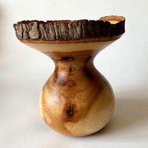 Puede incluir: Un jarrón de madera torneada con una base bulbosa y un borde ancho y acampanado. El exterior muestra una veta de madera de color marrón claro pulida con nudos visibles y un borde de corteza natural oscuro y texturizado en la parte superior.