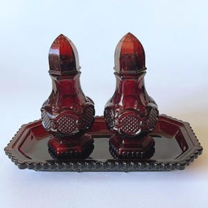 May include: A pair of deep red glass salt and pepper shakers with a textured, faceted pattern on the body and a matching rectangular glass tray with a scalloped edge. The shakers feature pointed, perforated lids and a hexagonal base design.