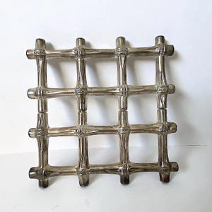 Peut inclure: Un dessous de plat carré composé de tiges métalliques argentées disposées en grille. Les tiges présentent un design texturé et segmenté imitant l'apparence de tiges de bambou, avec de petites bandes décoratives à chaque intersection.