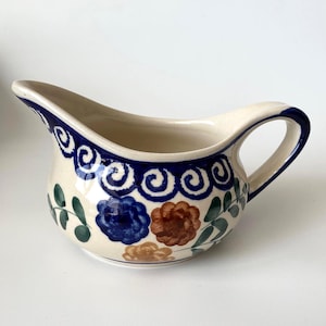 May include: Cream ceramic gravy boat with a blue, brown, and green floral pattern. The rim and handle are outlined in dark blue. The gravy boat has a spout for pouring and a curved handle.
