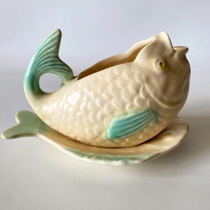 以下が含まれることがあります： クリーム色のセラミック製の魚の形をしたプランターで、ターコイズのアクセントが付いています。 プランターは、お揃いの魚の形をした皿の上に置かれています。 魚は、テクスチャ加工された鱗模様、ターコイズのひれ、側面にターコイズのアクセントがあります。