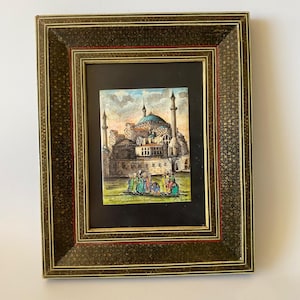 May include: A rectangular framed artwork featuring a painting of a mosque with a blue dome and minarets, with a group of figures in the foreground. The frame is dark brown with a gold-colored star pattern and thin red and gold inner borders.