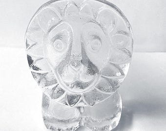 May include: A clear glass figurine shaped like a lion with a stylized mane. The object features a smooth, transparent surface with molded facial details including eyes, a nose, and a mouth, set within a circular frame representing the lion's head.