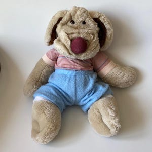 May include: A plush dog toy with a tan body, brown ears, and a red nose. It wears a red and white striped shirt and light blue shorts. The toy is soft and cuddly, perfect for children.