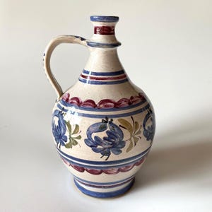 Op de afbeelding: Een keramische kan met een crèmekleurige basis, voorzien van handgeschilderde horizontale banden in blauw en bordeauxrood. Het lichaam is versierd met blauwe bloemmotieven en groene bladaccenten. Het heeft één gebogen handvat en een smalle hals.