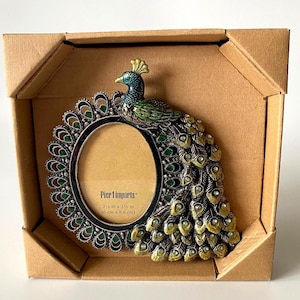May include: An oval picture frame shaped like a peacock with a metallic finish, featuring green and gold accents and embedded crystals. The frame displays the text Pier 1 imports and the dimensions 6 cm x 8.8 cm, housed in a brown box.