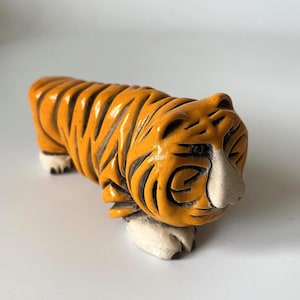 May include: A small, orange tiger figurine with black stripes and white paws. The tiger is carved with detailed lines and has a white nose. The figurine is a decorative item.
