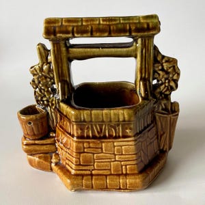 Può includere: Un vaso in ceramica smaltata marrone a forma di pozzo dei desideri, con base in mattoni e traversa superiore. La parte anteriore presenta il testo in rilievo A WISH, affiancato da dettagli floreali scolpiti e un piccolo secchio laterale.