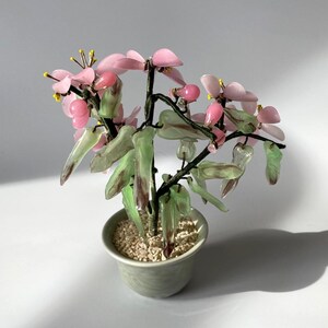 May include: A decorative miniature tree in a small, pale green ceramic pot. The tree features pink flower-shaped stones, green leaf-shaped stones, and small yellow accents. The pot is filled with small, light-coloured pebbles.
