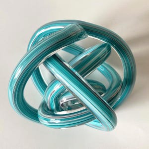 May include: A decorative glass sculpture formed from a continuous, knotted loop of clear glass tubing. The glass features internal parallel stripes in shades of teal, turquoise, and white, creating a swirling, multi-layered effect throughout the object.