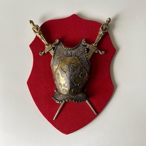 May include: A decorative wall plaque featuring a red felt shield-shaped backing. Mounted on the front is a metallic gold-toned breastplate with an embossed double-headed eagle emblem, flanked by two crossed swords with ornate, figure-topped hilts.
