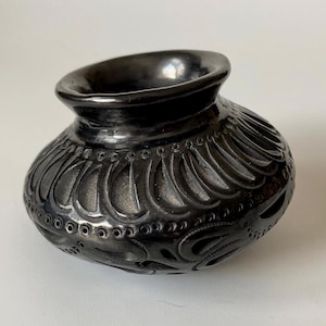 May include: A small, black ceramic pot with a rounded body and a flared rim. The pot has intricate carved designs, including teardrop shapes and small circular perforations. The dark colour and detailed carvings give it a handcrafted appearance.