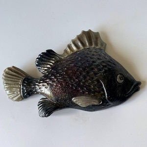 May include: A decorative fish figurine featuring a dark, textured body with metallic bronze and black tones. The fish has lighter, muted gold-coloured fins and tail, along with a single visible eye, showcasing a detailed scale pattern across its surface.