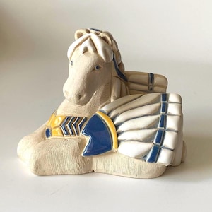 May include: A cream-coloured ceramic horse figurine in a seated position, with blue and yellow accents. The horse has a detailed mane and decorative elements on its chest and side, with a stylized wing-like design.