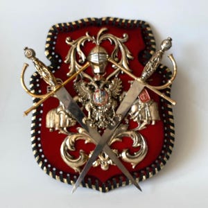 May include: A decorative shield with a red background, gold accents, and two crossed swords. The shield features an ornate design with a central emblem and a black and gold border. The swords have gold handles and silver blades.