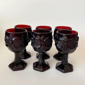 Può includere: Un set di sei calici in vetro rosso scuro caratterizzati da un motivo geometrico in rilievo sulla coppa, uno stelo corto e una base esagonale. Ogni calice presenta una superficie testurizzata con motivi a rombi e cerchi ben definiti.