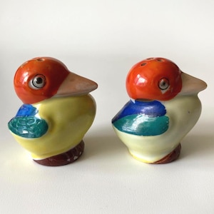 May include: A pair of ceramic bird-shaped salt and pepper shakers. Each bird features a bright orange head, a pale beige beak, a yellow body, and blue and green wings. The tops of the heads have small holes for dispensing seasoning.