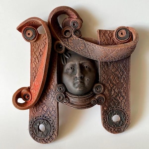 May include: A terracotta-coloured ceramic wall sculpture featuring a central dark grey human face surrounded by intricate, lace-like embossed patterns. The structure includes curved, scroll-like elements and two circular cutouts at the base.