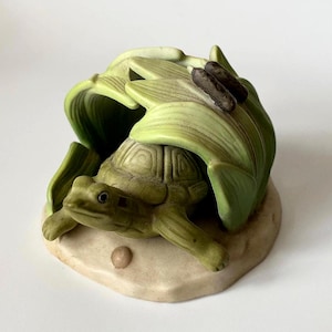 Może przedstawiać: Ceramiczna figurka żółwia w odcieniach zieleni i beżu. Żółw jest osadzony pod stylizowanymi zielonymi liśćmi z brązowymi akcentami. Figurka stoi na beżowej podstawie, tworząc dekoracyjny element inspirowany naturą.