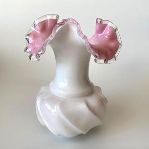 Può includere: Un vaso di vetro bianco con un bordo rosa a balze. Il vaso ha una base bulbosa con un motivo a spirale. Il bordo è trasparente con un interno rosa, creando un delicato contrasto. Un oggetto decorativo per la casa.