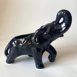 May include: A glossy black ceramic elephant ornament with its trunk raised. The elephant has a planter opening on its back and is standing on four legs. The ornament has a smooth, reflective surface and white accents on the feet.