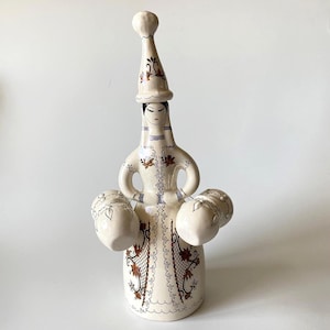 May include: A cream-colored ceramic decanter shaped like a figure wearing a tall pointed hat. The figure features painted brown and black floral patterns, a stylized face, and two small attached vessels on either side of the waist area.