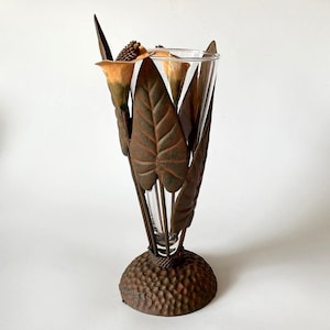 Peut inclure: Un vase en verre transparent inséré dans une structure métallique sombre et texturée, ornée de feuilles sculptées et d'éléments en forme de fleurs de lys. La base circulaire présente une surface martelée et le métal a une finition bronze vieilli.