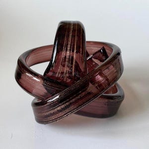 May include: A decorative glass sculpture in shades of purple and brown. The art glass piece is in a knotted design with gold-colored streaks. The sculpture is a unique home decor item.