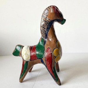 May include: A hand-painted horse figurine featuring a brown base color with vibrant accents in red, green, yellow, and white. The horse has a stylized mane with dark blue stripes and a large yellow flower design on its side, standing on four legs.