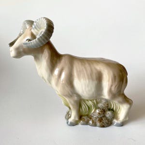 Puede incluir: Una figura de cerámica de un carnero de las Rocosas con acabado brillante. El animal presenta tonos crema, beige y marrón claro, con grandes cuernos curvados de color gris azulado. Se apoya sobre una base texturizada con detalles verdes y marrones.