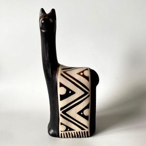 May include: A dark brown llama figurine with a long neck and pointed ears. The body features a cream-colored saddle blanket decorated with black geometric patterns, including triangles and circles, and a fringe detail at the bottom edge.