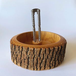 May include: A round wooden bowl with a natural bark exterior and a smooth, light brown interior. A silver-toned metal nutcracker is positioned vertically in the center of the bowl, attached to a small wooden base within the bowl's interior.