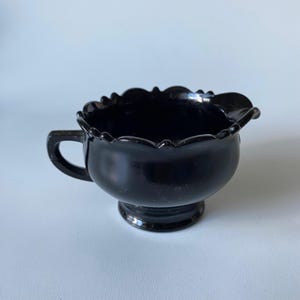 以下が含まれることがあります： 光沢のある黒いガラス製の小さなクリーマーピッチャー。縁はスカラップ状で、側面に湾曲したハンドルと小さな注ぎ口が付いています。容器は円形の台座の上にあり、外側全体が滑らかで光を反射する表面になっています。