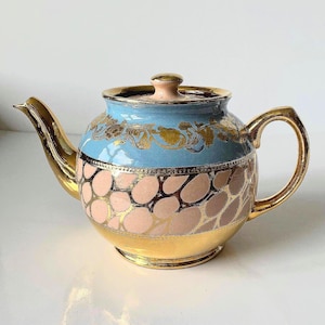 Peut inclure: Une théière en céramique avec une partie supérieure bleu clair ornée de motifs floraux dorés, une partie inférieure rose avec un motif de galets bordés d'or, ainsi qu'une base, un bec, une anse et un bouton de couvercle dorés.