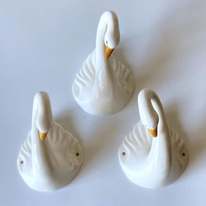 May include: Three white ceramic swan wall hooks with glossy finishes. Each swan features a curved neck, a yellow beak, and moulded wing details. The two lower hooks include visible mounting holes on the sides for secure wall attachment.