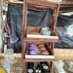 May include: A three-tiered wooden display stand filled with various handmade soaps and beauty products. The soaps are shaped like roses and other forms, in shades of purple and pink. Other products include small jars and bottles.