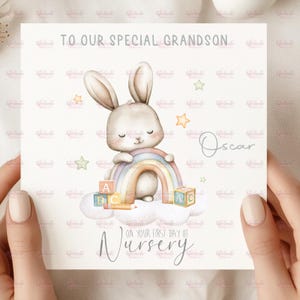 May include: A square greeting card featuring a watercolour illustration of a light brown bunny sitting on a white cloud with a pastel rainbow, alphabet blocks, and stars. The text reads TO OUR SPECIAL GRANDSON, Oscar, ON YOUR FIRST DAY OF NURSERY.