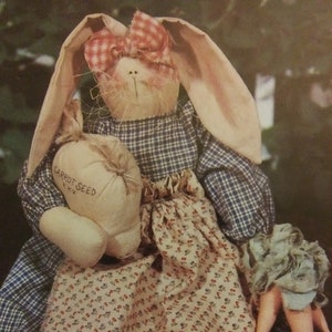 May include: A handmade fabric bunny doll wearing a blue and white plaid dress with a floral print apron. The bunny is holding a white fabric bag with the text "Carrot Seed" and has a carrot in its hand.