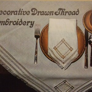 May include: A white linen tablecloth with a decorative drawn thread embroidery design. The tablecloth has a fringed edge and is set with a brown plate, a white napkin, a fork, a knife, and a spoon. The napkin has a diamond-shaped embroidery design in the center.