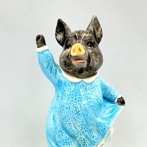 Peut inclure: Une figurine en porcelaine d'un cochon portant une robe bleue et debout sur une base verte. Le cochon lève sa main droite et a une expression joyeuse sur son visage. La figurine est une boîte à musique.