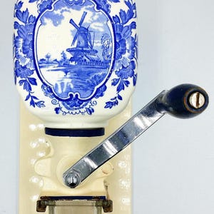 Op de afbeelding: Antieke keramische koffiemolen met een blauw-wit windmolenontwerp. De molen heeft een crèmekleurige basis, een metalen slinger en een kleine glazen container. De bovenkant is donkerblauw en de tekst "BendisCollectibles" is zichtbaar.