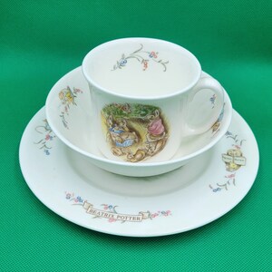Könnte beinhalten: Ein weißes Porzellan-Teeservice bestehend aus Tasse, Untertasse und Teller mit einer Illustration von Beatrix Potter mit Peter Rabbit und seinen Freunden. Das Set hat ein zartes Blumenmuster und die Worte "Beatrix Potter" auf dem Teller.